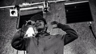 Black and white photo of person wearing a hoodie and black cap, rapping into a mic